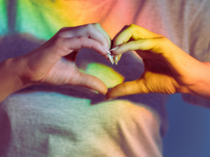 Image of person holding hands in a heart shape with a rainbow background